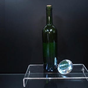 Pode incluir: Uma garrafa de vinho de vidro verde escuro com rolha de corti&ccedil;a est&aacute; ao lado de um peso de papel de vidro transparente facetado em forma de diamante. Ambos os itens est&atilde;o posicionados sobre um suporte de acr&iacute;lico transparente.