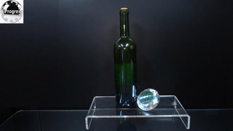 Pode incluir: Uma garrafa de vinho de vidro verde escuro com rolha de corti&ccedil;a est&aacute; ao lado de um peso de papel de vidro transparente facetado em forma de diamante. Ambos os itens est&atilde;o posicionados sobre um suporte de acr&iacute;lico transparente.