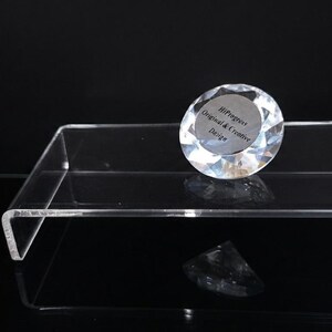 Pode incluir: Um peso de papel de vidro transparente facetado em forma de diamante sobre um suporte de exibi&ccedil;&atilde;o retangular de acr&iacute;lico transparente. O peso de papel apresenta um centro circular prateado com o texto HiProgress Original & Creative Design.
