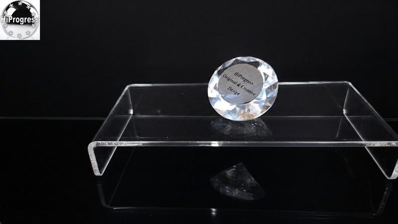 Pode incluir: Um peso de papel de vidro transparente facetado em forma de diamante sobre um suporte de exibi&ccedil;&atilde;o retangular de acr&iacute;lico transparente. O peso de papel apresenta um centro circular prateado com o texto HiProgress Original & Creative Design.