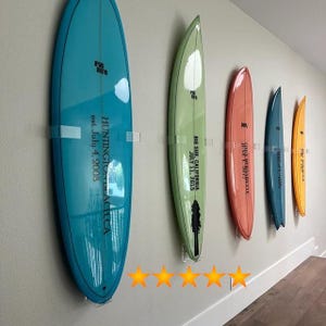 May include: Five surfboards are mounted on a wall using clear acrylic surfboard racks. The surfboards are all different colors and have different text on them. The text on the surfboards reads: "HUNTINGTON BEACH CA est. July 4, 2008", "BIG SUR, CALIFORNIA JULY 11, 2015", "SHIVA PENINSULA", "JUNK" and "PIG IN THE PYGMY".