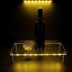 May include: A clear rectangular acrylic display riser with integrated yellow LED lighting along the edges, supporting a dark glass wine bottle and a small circular white tag with black text, illuminated by a yellow light source from above.