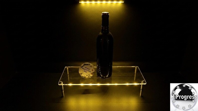 May include: A clear rectangular acrylic display riser with integrated yellow LED lighting along the edges, supporting a dark glass wine bottle and a small circular white tag with black text, illuminated by a yellow light source from above.