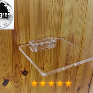 Könnte beinhalten: Ein klares Acrylregal mit quadratischer Form und abgerundeten Kanten. Das Regal ist mit einer klaren Acrylhalterung an einer Holzwand befestigt. Das Regal ist für die Aufbewahrung kleiner Gegenstände konzipiert.
