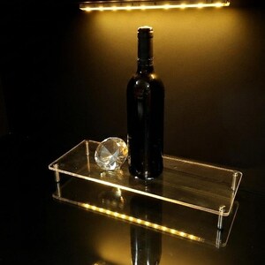 May include: A rectangular clear acrylic display riser with four metal legs, featuring an integrated LED light strip along the front edge. A dark glass wine bottle and a faceted clear glass diamond-shaped paperweight sit on the flat surface.