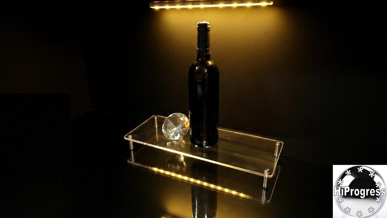 May include: A rectangular clear acrylic display riser with four metal legs, featuring an integrated LED light strip along the front edge. A dark glass wine bottle and a faceted clear glass diamond-shaped paperweight sit on the flat surface.