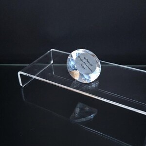 Pode incluir: Um suporte de exibi&ccedil;&atilde;o retangular de acr&iacute;lico transparente com perfil em U. Na superf&iacute;cie repousa um peso de papel de vidro transparente facetado em forma de diamante com um adesivo circular com o texto HiProgress Original & Creative Design.