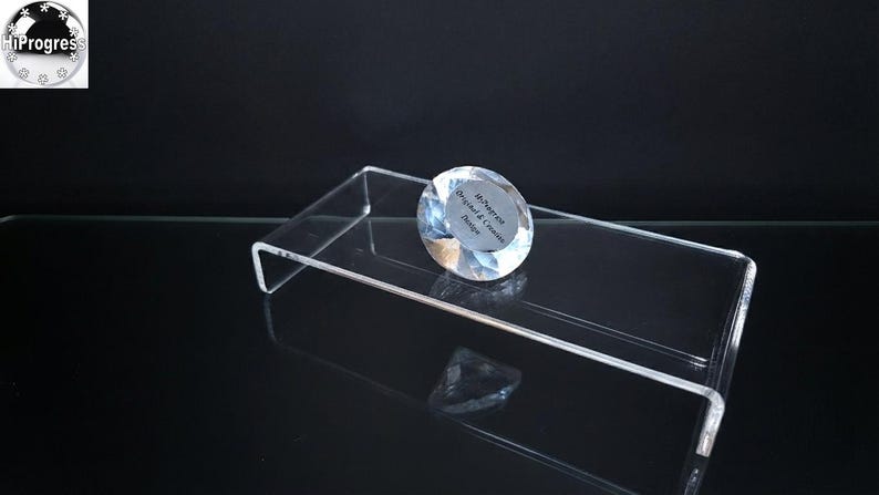 Pode incluir: Um suporte de exibi&ccedil;&atilde;o retangular de acr&iacute;lico transparente com perfil em U. Na superf&iacute;cie repousa um peso de papel de vidro transparente facetado em forma de diamante com um adesivo circular com o texto HiProgress Original & Creative Design.