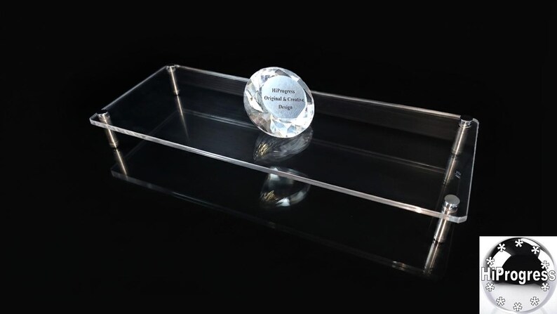 May include: A rectangular clear acrylic display riser with four silver metal cylindrical legs. Resting on the surface is a faceted clear glass diamond-shaped paperweight featuring the text HiProgress Original & Creative Design.
