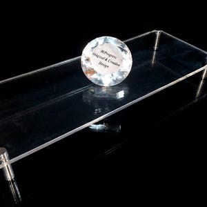 May include: A rectangular clear acrylic display riser with four silver-toned metal cylindrical legs. A round, faceted, clear glass paperweight sits on the surface, featuring the printed text HiProgress Original & Creative Design.