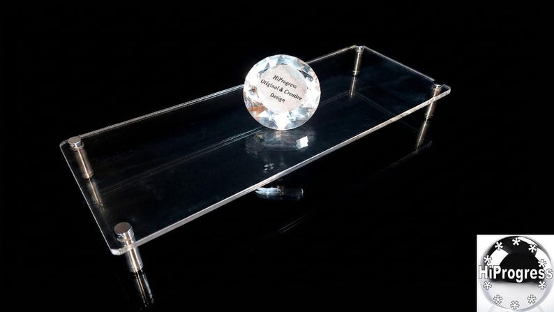 May include: A rectangular clear acrylic display riser with four silver-toned metal cylindrical legs. A round, faceted, clear glass paperweight sits on the surface, featuring the printed text HiProgress Original & Creative Design.