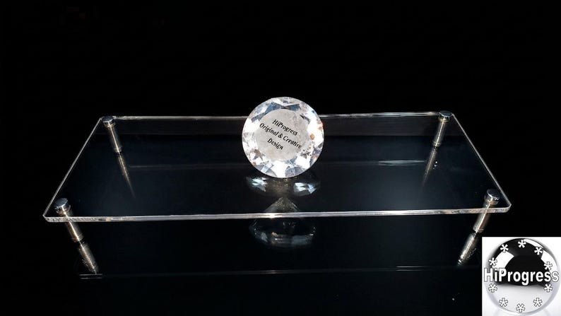 May include: A rectangular clear acrylic display stand supported by four silver-toned metal cylindrical legs. A circular, faceted clear crystal-like object sits on the center of the platform, featuring the text HiProgress Original & Creative Design.