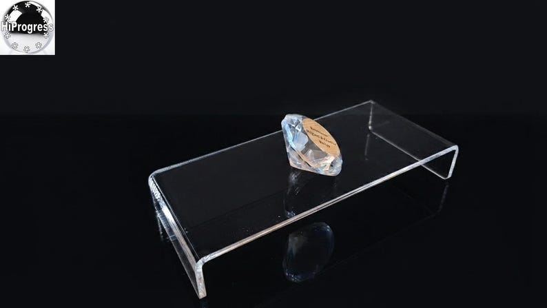 Pode incluir: Um suporte expositor retangular de acr&iacute;lico transparente com design em forma de U, apresentando um peso de papel de vidro facetado em forma de diamante sobre a superf&iacute;cie. O peso de papel tem uma incrusta&ccedil;&atilde;o circular dourada com texto.