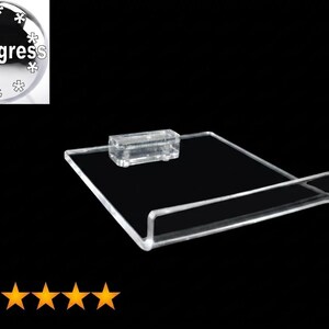 May include: A clear acrylic display stand featuring a flat rectangular base with a raised front edge and a small rectangular block attachment on the back. The design is transparent and minimalist, intended for showcasing items on a flat surface.
