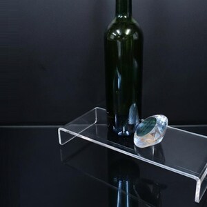 Pode incluir: Um expositor retangular de acr&iacute;lico transparente com design em forma de U. Uma garrafa de vinho de vidro verde escuro est&aacute; na vertical sobre a superf&iacute;cie, acompanhada por um pequeno peso de papel de vidro transparente facetado.