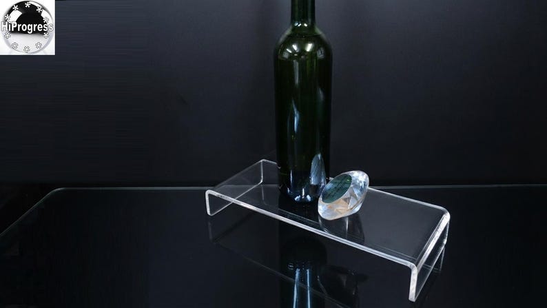 Pode incluir: Um expositor retangular de acr&iacute;lico transparente com design em forma de U. Uma garrafa de vinho de vidro verde escuro est&aacute; na vertical sobre a superf&iacute;cie, acompanhada por um pequeno peso de papel de vidro transparente facetado.