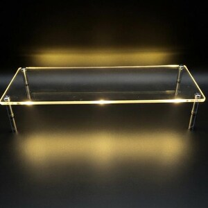 May include: A rectangular clear acrylic display riser featuring a smooth flat surface supported by four cylindrical metal legs. The edges of the transparent platform emit a soft warm yellow glow, creating a luminous effect against the dark background.