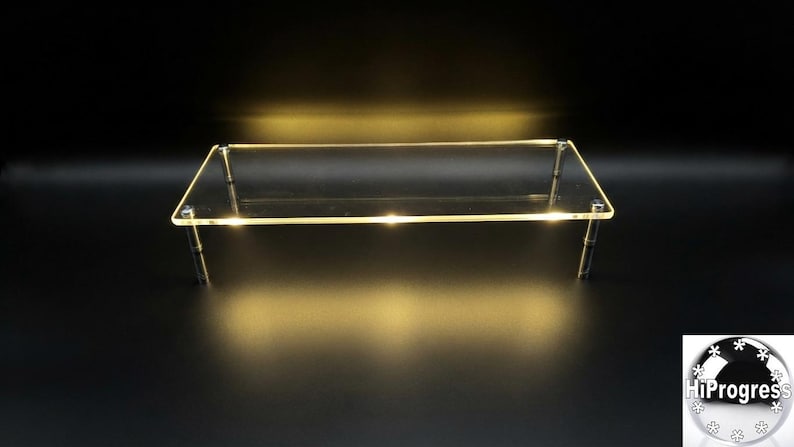 May include: A rectangular clear acrylic display riser featuring a smooth flat surface supported by four cylindrical metal legs. The edges of the transparent platform emit a soft warm yellow glow, creating a luminous effect against the dark background.
