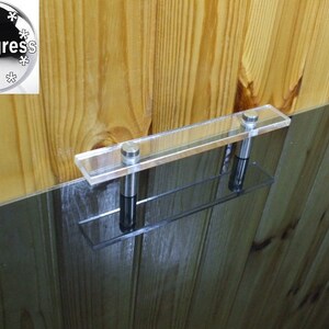 May include: A clear acrylic shelf with two stainless steel supports, mounted on a wooden wall. The shelf is rectangular and appears to be designed for displaying items. The wood has a natural grain pattern.