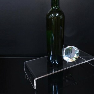 Pode incluir: Um expositor retangular de acr&iacute;lico transparente com design em forma de U. Uma garrafa de vidro verde escuro est&aacute; na vertical no centro do expositor, acompanhada por um pequeno objeto decorativo facetado de cristal transparente ao lado.