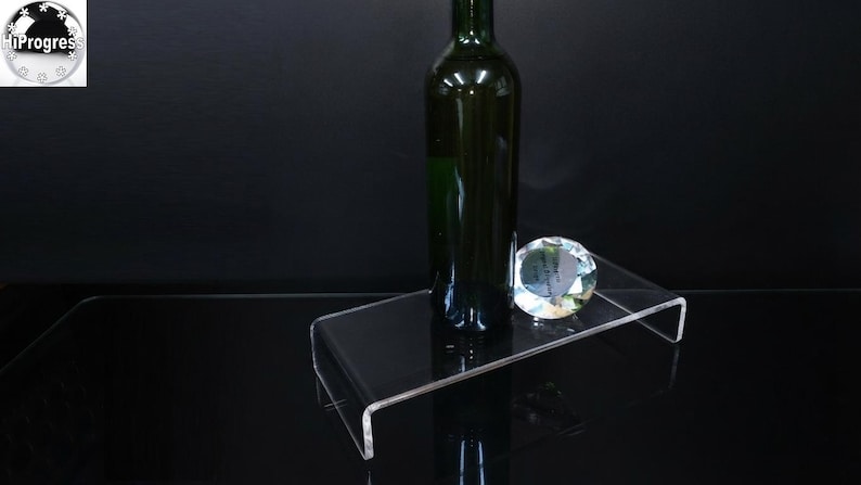 Pode incluir: Um expositor retangular de acr&iacute;lico transparente com design em forma de U. Uma garrafa de vidro verde escuro est&aacute; na vertical no centro do expositor, acompanhada por um pequeno objeto decorativo facetado de cristal transparente ao lado.