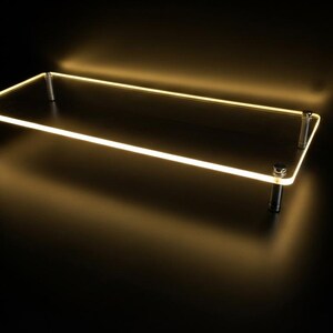 May include: A rectangular clear acrylic display riser featuring a glowing warm yellow LED edge light. The platform is supported by four cylindrical metal legs positioned at each corner, creating a floating effect against a dark background.