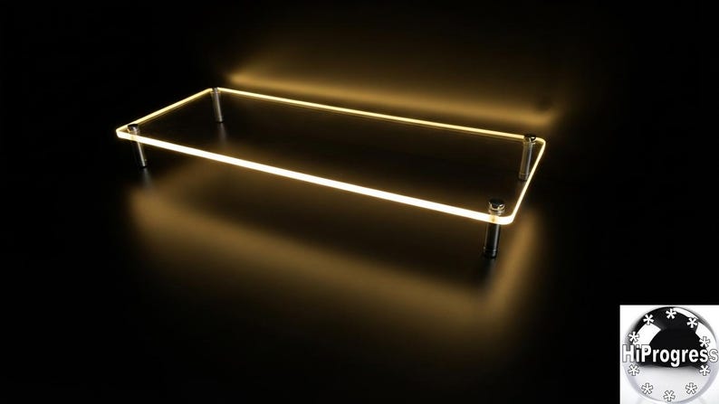 May include: A rectangular clear acrylic display riser featuring a glowing warm yellow LED edge light. The platform is supported by four cylindrical metal legs positioned at each corner, creating a floating effect against a dark background.