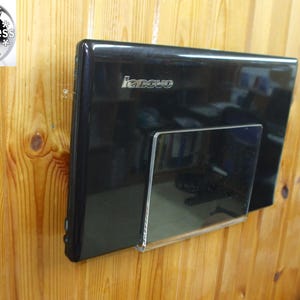 Könnte beinhalten: Ein schwarzer Lenovo-Laptop, montiert auf einem transparenten Acrylhalter an einer Holzwand. Der Laptop ist vertikal positioniert, mit dem Lenovo-Logo sichtbar. Die Wand hat ein natürliches Holzmaserungsmuster.