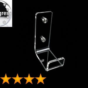 Puede incluir: Un soporte de pared en forma de L hecho de acrílico transparente, que cuenta con dos fijaciones circulares de metal plateado. El expositor tiene un panel trasero rectangular y una base plana diseñada para sostener objetos contra la pared.