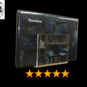 May include: A black Lenovo laptop lid featuring a glossy finish and the silver Lenovo logo in the upper left corner. A clear acrylic wall-mount holder is attached to the front surface, and five orange stars are positioned below the laptop.