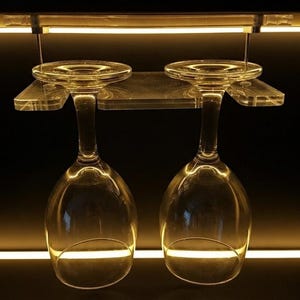 May include: Two clear glass wine glasses hanging upside down from a transparent acrylic rack. The rack is mounted under a shelf illuminated by a warm horizontal LED light strip. The text HiProgress is visible in the bottom right corner.