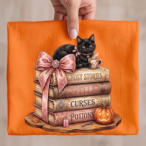 Op de afbeelding: Een oranje t-shirt met een afbeelding van een zwarte kat met een roze strik en halsband, zittend op een stapel van vier boeken met de titels Ghost Stories, Spells, Curses en Potions. Een grote roze strik, een dampende mok, een pompoen, een flesje en een sleutel.
