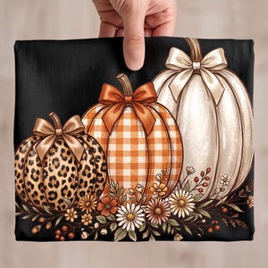 May include: A black fabric featuring three pumpkins with bows. The left pumpkin has a leopard print, the centre one has an orange and white gingham pattern, and the right one is white. The base is decorated with white daisies and small brown berries.
