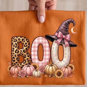 Op de afbeelding: Een oranje t-shirt met de tekst BOO in grote letters. De B heeft een luipaardprint, de O een roze gingham-patroon en de laatste O is wit met parels en een zwarte heksenhoed met een roze strik. Pompoenen, zonnebloemen en rozen staan eronder.