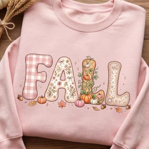May include: A light pink crewneck sweatshirt featuring the word FALL in large decorative letters. The F has a pink gingham pattern, the A has a floral print, the L features pumpkin vines, and the second L is decorated with white pearls.