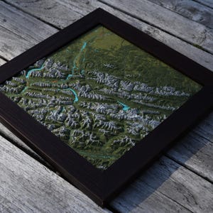 Pode incluir: Uma moldura quadrada de madeira castanho-escura contendo um mapa topográfico tridimensional. O mapa exibe cadeias montanhosas detalhadas com picos cinzentos, vales verdes e rios azul-turquesa brilhantes pelo terreno.