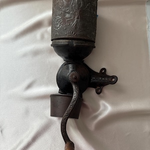 May include: A wall-mounted cast iron coffee grinder with a dark, weathered finish. The cylindrical hopper features an embossed floral pattern. A curved metal handle with a wooden knob extends from the side, and the word ROYAL is cast into the mounting bracket.