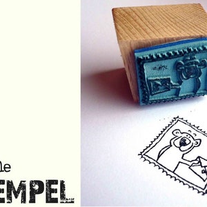 Könnte beinhalten: Ein Holzstempel mit einer blauen Gummistempelfläche, die einen Cartoon-Bären mit einem Umschlag zeigt. Der Stempel ist auf eine weiße Oberfläche gedrückt und erzeugt ein Schwarz-Weiß-Bild des Bären. Der Text "freche STEMPEL" ist links aufgedruckt.