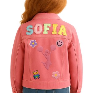 May include: Pink denim jacket with the name "SOFIA" in colorful letters on the back. The jacket features patches of a cheerleader, flowers, a smiley face, and a bear. The jacket is a children's apparel item.