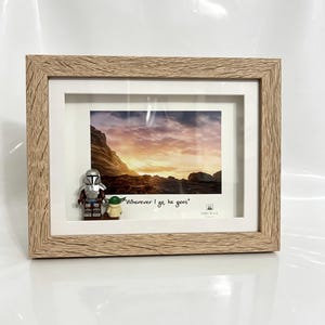 May include: A framed print featuring a sunset landscape with Lego figures of The Mandalorian and Grogu. The frame is light brown with a textured finish. The print includes the text "Wherever I go, he goes."