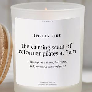 May include: A white frosted glass candle with a wooden lid. The candle has a label that reads "Smells Like the calming scent of reformer pilates at 7am". The candle is hand-poured in the USA.