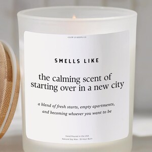 May include: A frosted glass candle with a wooden lid. The candle has a white label with the text "SMELLS LIKE the calming scent of starting over in a new city". The candle is lit.