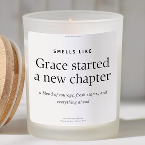 May include: A white frosted glass candle with a wooden lid. The candle has a label that reads "Smells Like Grace started a new chapter." The text below says "a blend of courage, fresh starts, and everything ahead."