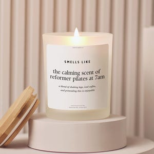 May include: A lit candle in a frosted glass jar with a wooden lid. The label reads "SMELLS LIKE the calming scent of reformer pilates at 7am." The candle sits on a white pedestal, with the lid resting nearby.