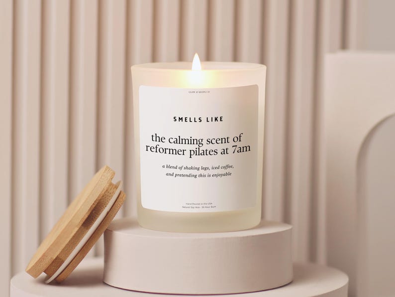 May include: A lit candle in a frosted glass jar with a wooden lid. The label reads "SMELLS LIKE the calming scent of reformer pilates at 7am." The candle sits on a white pedestal, with the lid resting nearby.