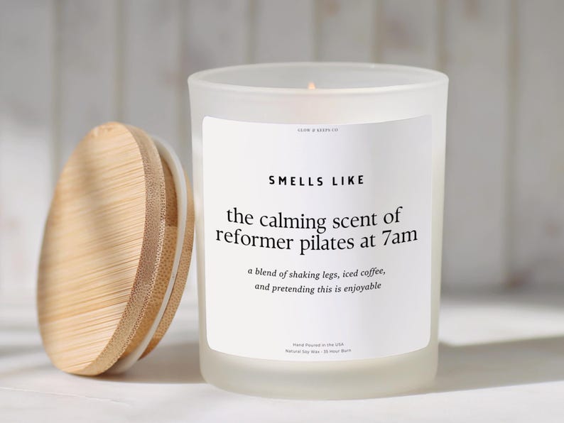 May include: A white frosted glass candle with a wooden lid. The candle has a label that reads "Smells Like the calming scent of reformer pilates at 7am". The candle is hand-poured in the USA.