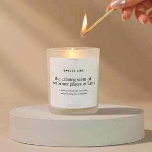 May include: A lit candle in a frosted glass jar on a white circular pedestal. The candle has a white label with the text "SMELLS LIKE the calming scent of reformer pilates at 7am". A match is held above the candle.