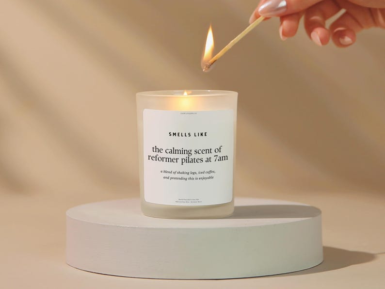 May include: A lit candle in a frosted glass jar on a white circular pedestal. The candle has a white label with the text "SMELLS LIKE the calming scent of reformer pilates at 7am". A match is held above the candle.