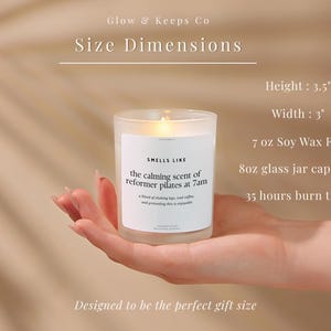 May include: A lit candle in a clear glass jar, held in a hand. The candle has a white label that reads "Smells Like the calming scent of reformer pilates at 7am." The candle is 3.5 inches tall and 3 inches wide, with a 7 oz soy wax fill and 8oz glass jar capacity.