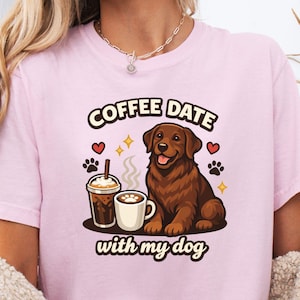 May include: A pale pink t-shirt with a brown dog illustration and the words "COFFEE DATE with my dog". The design includes coffee cups, hearts, and paw prints. The shirt is made of a soft fabric.